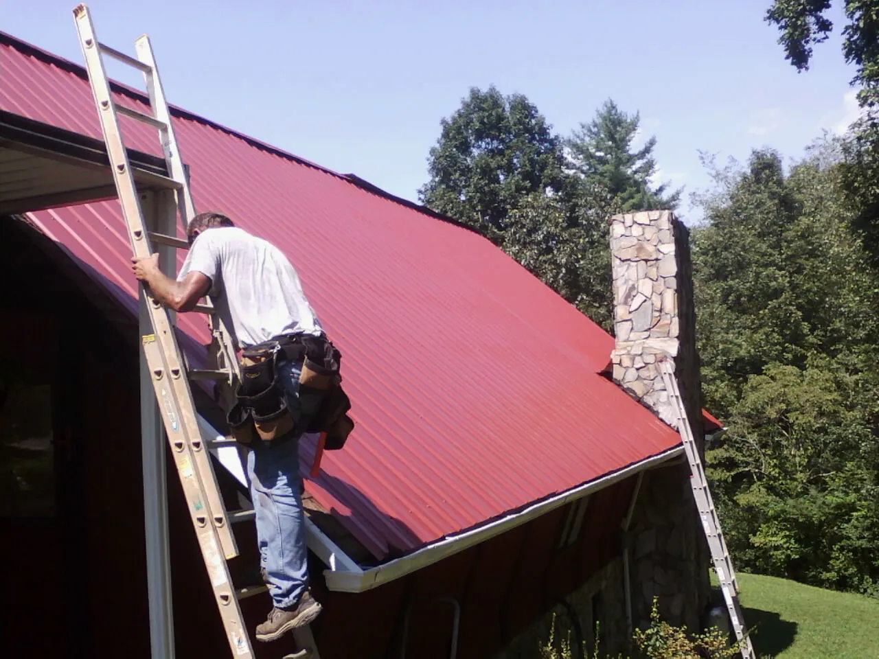 Roofer on a ladder beside a red metal roof and stone chimney — Soffit & Fascia Repair by Roofing Brothers in Armagh